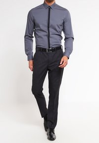 Camicia grigia con bottoni e striscia verticale nera a contrasto, abbinata a pantaloni scuri a trama e scarpe eleganti nere.