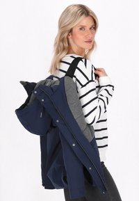 Námornícky modrá bunda so šedým interiérom, so zipsom na prednej strane a kapucňou. Modelka má na sebe pruhované tričko s dlhým rukávom a nesie bundu cez ramena.