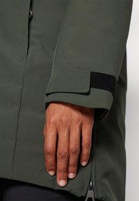 Zelená nepremokavá bunda s hladkým povrchom, so západkou na suchý zips na manžete a detailom zipsu na leme.