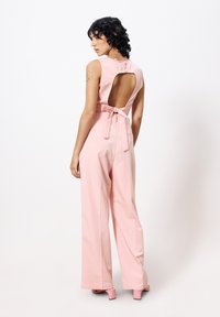 Rosa Jumpsuit mit offener Rückseite und Bindeband in der Taille. Verfügt über weite Beine und eine glatte Textur. Maßgeschneiderte Schultern und minimales Zubehör.