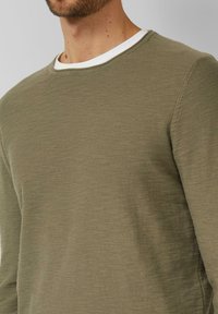 Mężczyzna ubrany w oliwkowozielony sweter z okrągłym dekoltem na białej koszulce z okrągłym kołnierzem, pokazany od ramion do połowy tułowia.