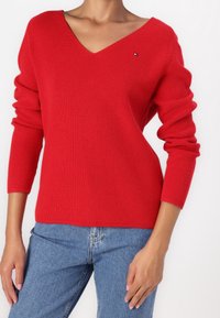 Maglione rosso a coste con scollo a V e maniche lunghe, con un piccolo dettaglio logo. Abbinato a jeans di denim blu.