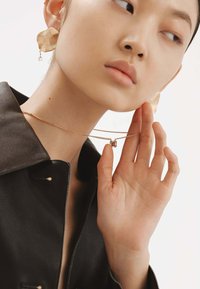 Pendientes con textura dorada y un collar de cadena dorada en capas con un pequeño colgante rectangular. El fondo es simple para centrar la atención en los accesorios.