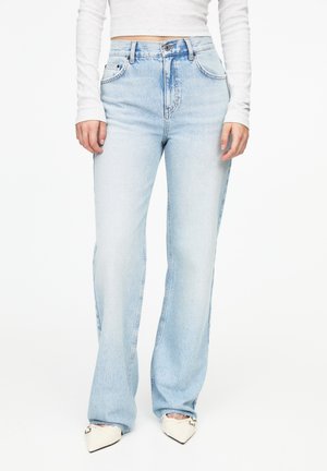 Femme portant un jean bleu clair taille haute à jambes droites, un top court blanc à manches longues et des chaussures blanches pointues avec des bouts noirs.