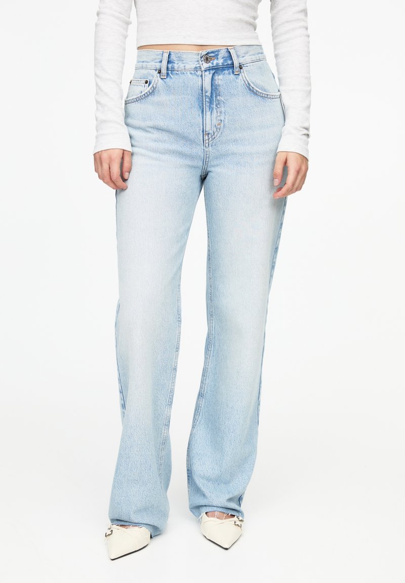 Femme portant un jean bleu clair taille haute à jambes droites, un top court blanc à manches longues et des chaussures blanches pointues avec des bouts noirs.