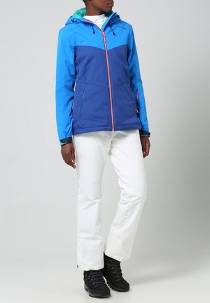 Veste isolante bleue et bleu sarcelle avec fermeture éclair à l'avant, fermeture éclair rose contrastante et poches, associée à un pantalon blanc et des bottes noires.