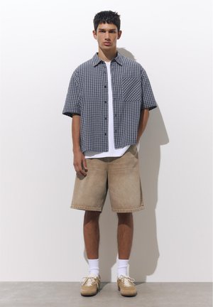 Tânăr stând lângă un perete alb, purtând o cămașă scurtă cu mâneci albastre în carouri, un tricou alb, pantaloni scurți bej, șosete albe și adidași bej.