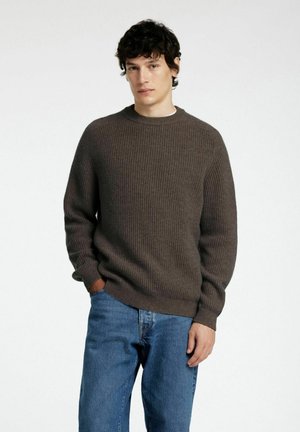 Brown ribbed knit sweater with a crew neckline. Paired with blue denim jeans, model poses with hands in pockets against a plain background.