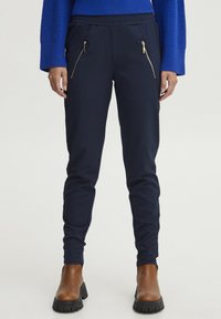 Marina blå tapered byxor med elastisk midja, dragkedjefickor och en slät textur, kombinerade med bruna ankelstövlar och en blå stickad tröja.