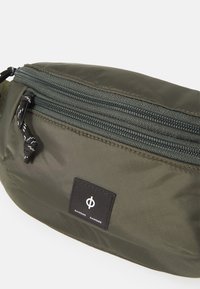 Marsupio in nylon verde oliva con due scomparti con zip, toppa con logo nera e finitura testurizzata. Presenta tirazip cordonati in bianco e nero.