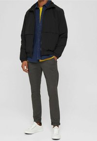 Giacca piumino nera con zip frontale, indossata sopra una camicia button-up blu e una maglietta gialla, abbinata a pantaloni grigi e sneaker bianche.
