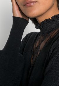 Haut noir à col montant avec col à volants, superposé sur une dentelle transparente. Manches longues ajustées avec texture côtelée, dévoilant un subtil effet drapé.