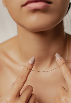 Goldkette mit kleinen, runden Perlen, gehalten zwischen zwei Fingern. Der Hintergrund ist neutral und lenkt den Fokus auf das Design und die Textur des Schmucks.