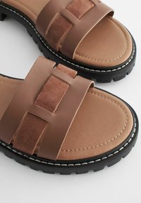 Braune Rutschsandalen mit doppelt strukturierten Riemen, einem glatten Kunstleder- und einem weichen Wildlederriemen. Dicke schwarze Gummisohle mit weißer Naht.