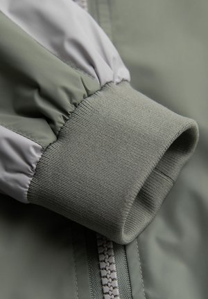 Detail van de mouw van een groene jas met elastische manchetten en een gladde, glanzende stof. Bevat een contrasterende grijze accent bij de schouder.