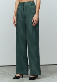 Groene gestreepte broek met een hoge taille, brede pijpen en plooien. De stof is glad, accentueert met een riem en is gecombineerd met zwarte hakken.
