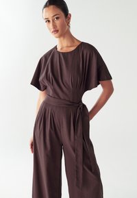 Brun jumpsuit med lös passform, korta ärmar och midjebälte. Har veck och en slät, matt yta.