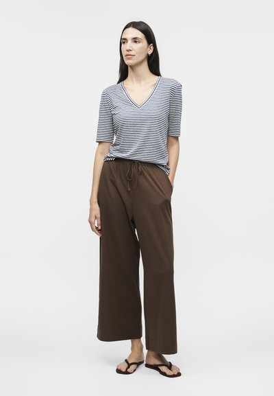 Γυναίκα με μακριά σκούρα μαλλιά, φορώντας ριγέ μπλούζα με V λαιμόκοψη και κοντά μανίκια, καφέ φαρδιά παντελόνια με κορδόνι, και μαύρα σαγιονάρες, όρθια.