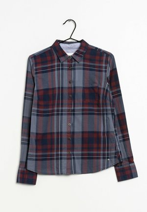 Camisa de manga larga con botones y patrón de cuadros en rojo, azul marino y gris colgada en una percha negra sobre fondo blanco.