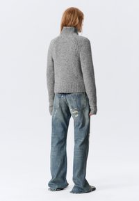 Γκρι πλεκτό πουλόβερ με ψηλό γιακά, ριμπ μανίκια και μακριά μανίκια, σε συνδυασμό με σκισμένα μπλε τζιν και μαύρα παπούτσια.