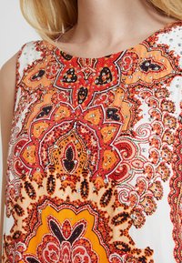 Ärmelloses Oberteil mit einem auffälligen orange-weißen Paisleymuster, ergänzt durch schwarze und rote Akzente, verziert mit silbernen Nieten für Textur.