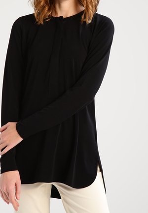 Vrouw die een zwart tuniek bovenstuk met lange mouwen en zijopeningen draagt, gecombineerd met crèmekleurige broek, tegen een effen witte achtergrond.