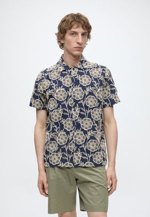 Jeune homme portant une chemise bleu marine à manches courtes avec une broderie florale beige et un short vert olive, debout devant un fond uni.