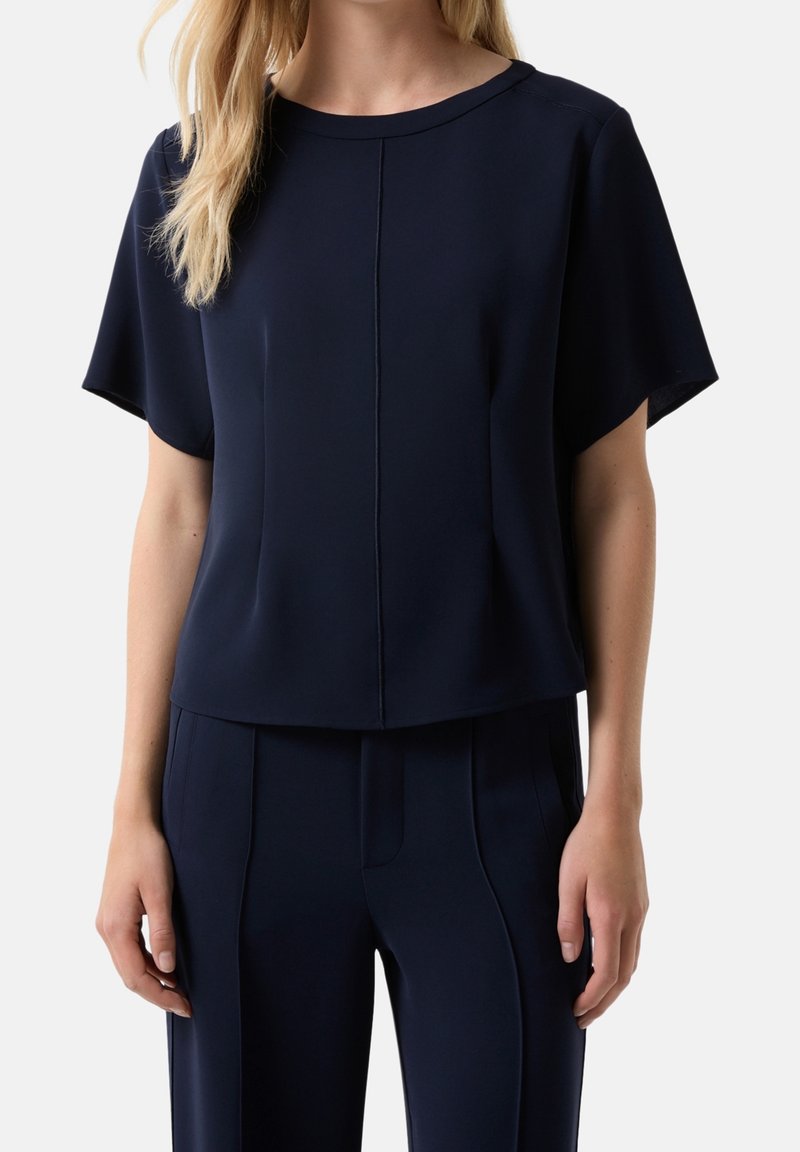 Femme portant un haut bleu marine à manches courtes avec un col rond et un pantalon bleu marine assorti avec des coutures devant.