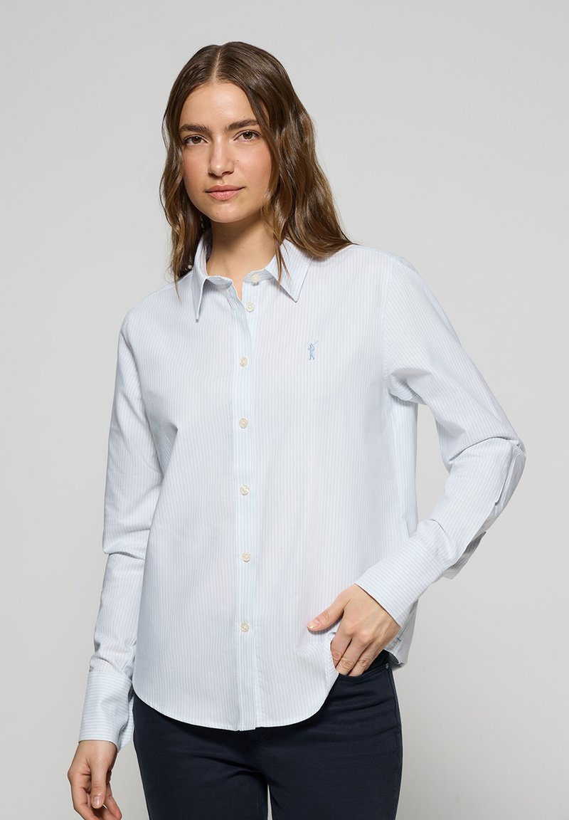 Ljusblå randig skjorta med knappar och långa ärmar, knappmanschetter och rundad nederkant. Har en liten broderad logotyp på vänster bröst.