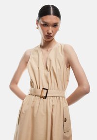 Beige mekko ilman hihoja, jossa on syvä V-pääntie, nyöritetty vyötärö ja vyö, jossa on suorakulmainen kultasolki. Mukana on kaksi sivutaskua.