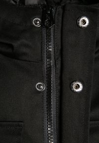 Musta takki, jossa on keskellä vetoketju, teksturoitu kangas, painonapit ja kestävä materiaali. Design korostaa kestävyyttä ja toiminnallisuutta.