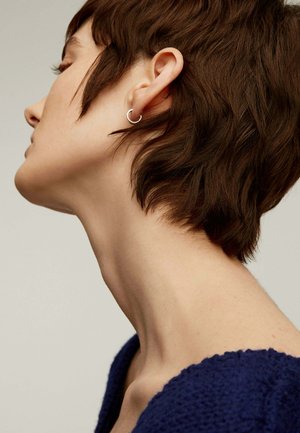 Pendiente de aro plateado llevado en un cabello corto y texturizado castaño. El cuello está en ángulo, revelando un tono de piel suave y claro contra un suéter azul marino.