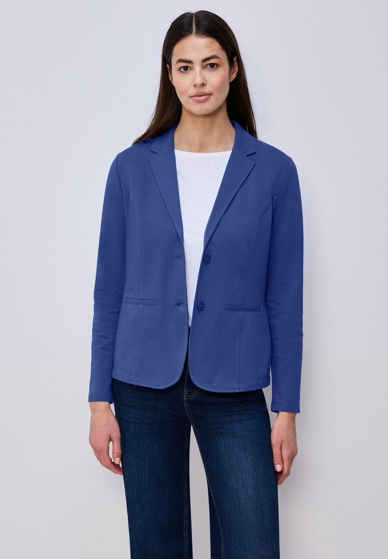 Femme aux cheveux raides et foncés portant un blazer bleu sur un haut blanc et un jean bleu foncé, debout devant un fond clair uni.