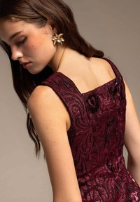 Vestido burdeos sin mangas con intrincados patrones florales de terciopelo; la modelo lleva un pendiente dorado con flores; tejido texturizado, escote cuadrado.