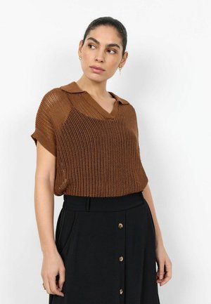 Žena u smeđoj pletenoj majici kratkih rukava sa ovratnikom i crnoj suknji visokog struka s prednjim gumbima, okreće se lijevo.