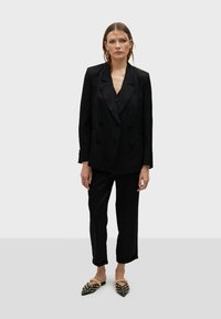 Černý dvouřadý blazer s texturovanou tkaninou, spárovaný s odpovídajícími kalhotami a černými slip-on botami se cvočky. Minimalistický design.