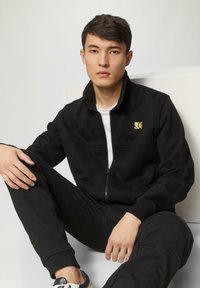 Schwarz zip-up Jacke mit hohem Kragen und kleinem goldenen Logo auf der Brust; kombiniert mit schwarzen Jogginghosen. Weicher Baumwollstoff, lässige Passform.