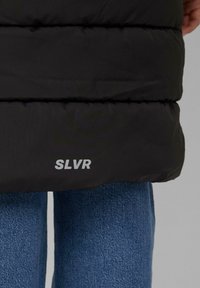 Schwarze Steppjacke mit einem ge quilteten Design; verfügt über ein silbernes "SLVR"-Logo am Saum. Getragen über blauen Jeans.