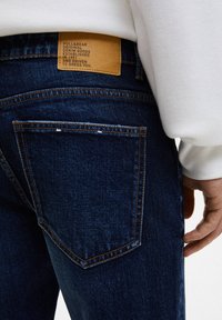 Tummansiniset farkut, joiden takatasku on näkyvissä, kontrastipistelyt ja ruskea nahkainen merkki tekstillä vyötärönauhasta.