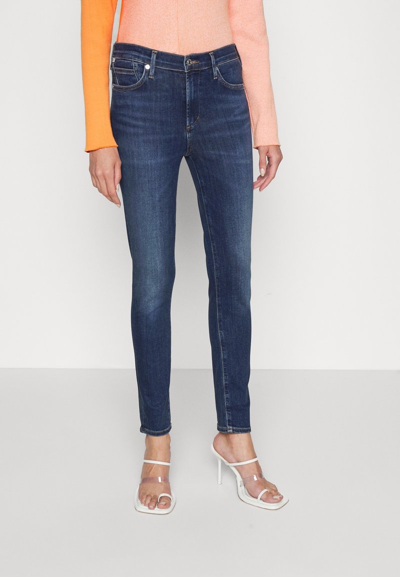 Slim-fit, mörkblå jeans med medelhög midja, fem-fickorsdesign och lätt blekning på låren; parat med klara högklackade sandaler.