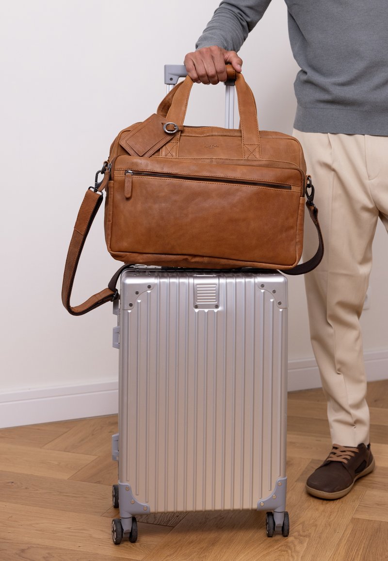 Rudas odinis kelioninis krepšys su petine juosta uždėtas ant sidabrinio, raukšlėto kietos dangos lagamino su pilkai ratais. Krepšys turi priekinį užtrauktuką.