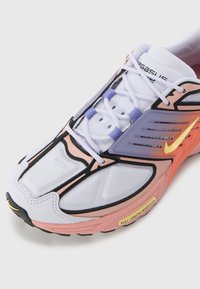 Fehér és rózsaszín sportcipő fekete kiegészítőkkel, hálós felsőrészzel, lila fűzőkkel, és hangsúlyos sárga Nike logóval; textúrált dizájn elemekkel rendelkezik.