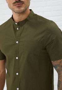 Short-sleeved olive green shirt in lightweight fabric, featuring a buttoned front, collar, and a single chest pocket with white stitching.