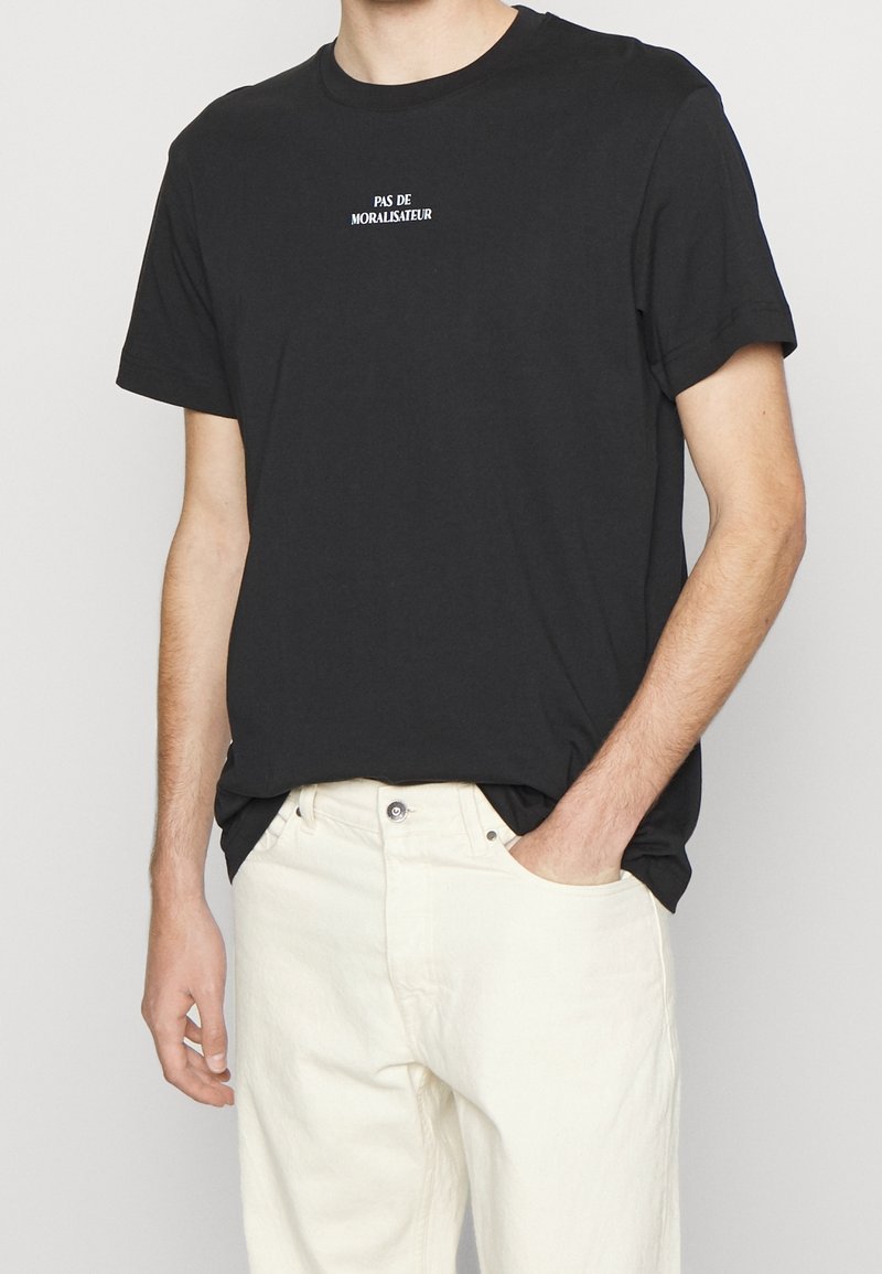 Czarna bawełniana koszulka z okrągłym dekoltem. Z przodu biała napis "PAS DE MORALISATEUR". Połączona z jasnymi dżinsami.