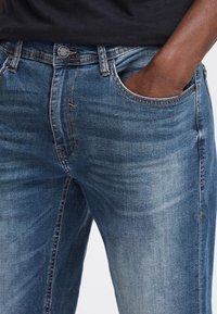 Jean en denim bleu pour homme avec une coupe classique, cinq poches et des coutures contrastées. Texture lisse avec un léger dégradé au niveau des cuisses.