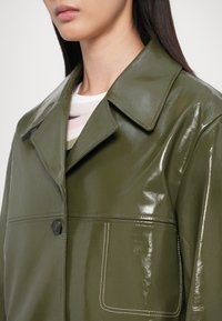 Groene glanzende leren jas met een puntkraag, sluiting met één knop en een voorzak die wordt benadrukt door witte stiksels.