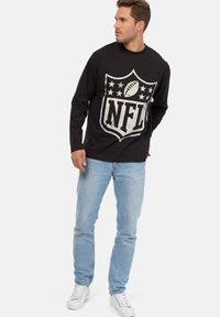 Maglietta nera a maniche lunghe con un grafico del logo NFL in stile usurato sul fronte, abbinata a jeans azzurri e sneaker bianche.