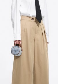 Geantă mini fabricată din material asemănător denimului, cu mânere din piele maro și închidere cu fermoar. Purtyată alături de pantaloni largi bej deschis și o cămașă albă.