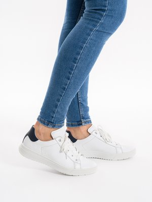 Personne portant des baskets en cuir blanc avec un détail noir au talon et un jean skinny bleu, debout sur une surface blanche.