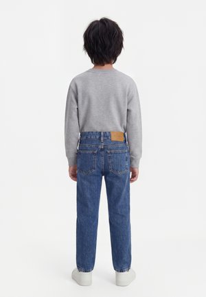 Niño con cabello oscuro de pie de espaldas, lleva una sudadera gris, vaqueros azules con bolsillos traseros y zapatillas blancas sobre un fondo blanco.